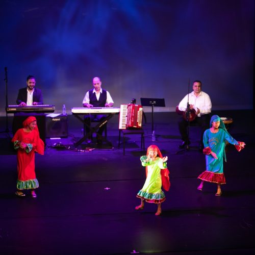 Three small children dance on a dark stage. Their brilliantly colored outfits of red, lime green and sapphire blue shine as they move.