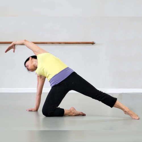 Woman with a yellow top and black yoga pants stretching in a studio with white walls and grey flooring.